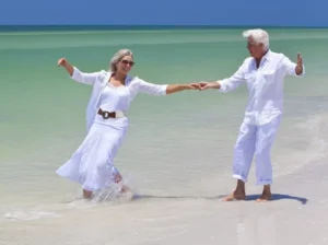 Icouple walking along the beach in shallow water, dressed in white doing a dance move as they enjoy their retirement in langebaan